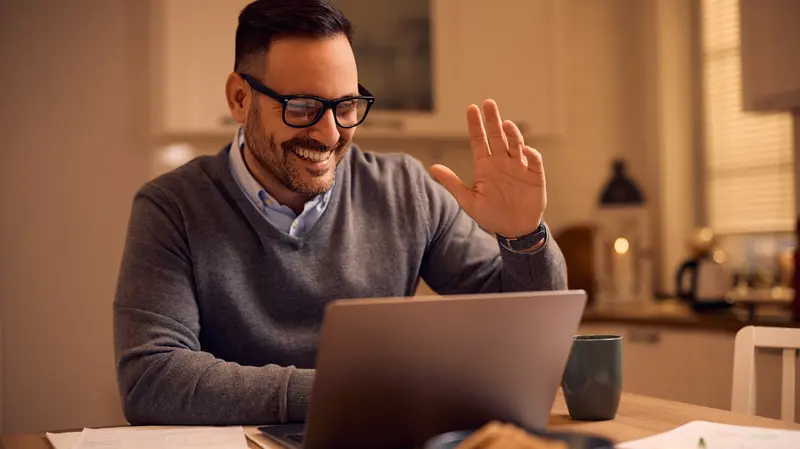 A man smiling and waving during a video call on his laptop at a kitchen table in a warmly lit home.