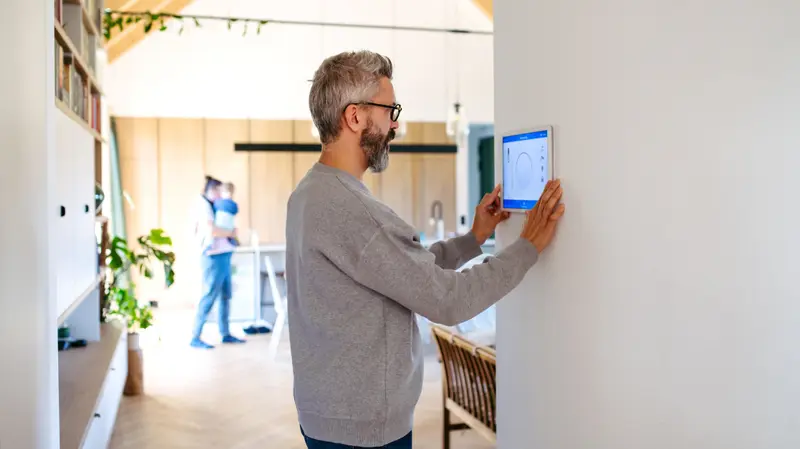 A man adjusting a smart home control panel mounted on the wall of a modern, open-plan home.