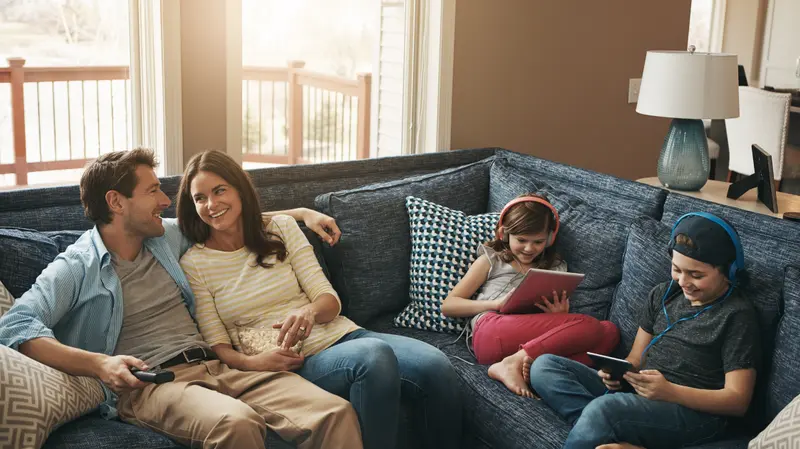 A family relaxing on the couch together, with two children using tablets and headphones while their parents watch TV in the living room.