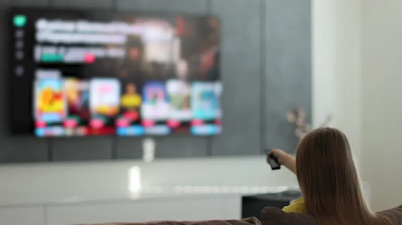 A woman pointing a remote at a large smart TV displaying a streaming service home screen in a modern living room.