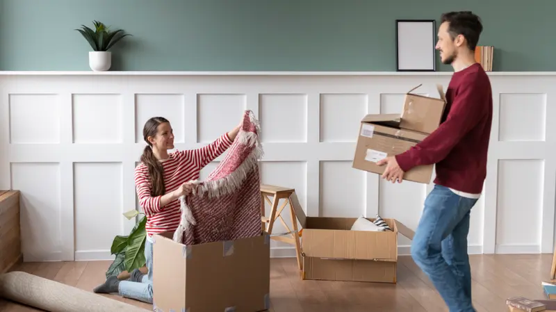 A couple unpacking moving boxes together in a bright, modern living room with wainscoting and warm wood floors.