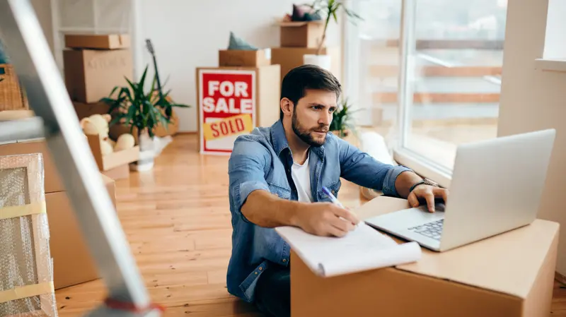 Man sitting on the floor of a home surrounded by moving boxes, using a laptop and taking notes, with a sold real estate sign visible in the background.