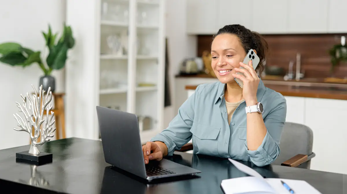 Woman sitting at a home desk, smiling while talking on her smartphone and typing on a laptop, with a bright kitchen in the background.