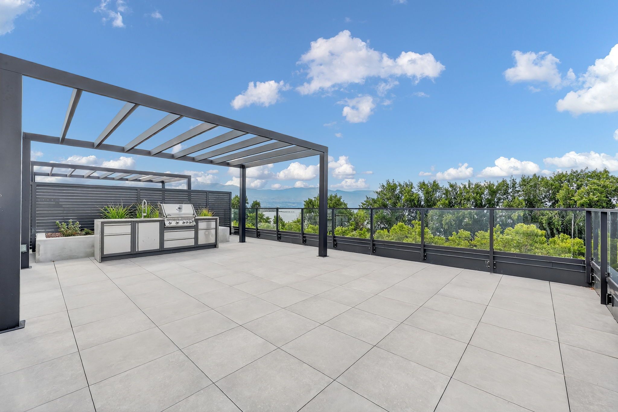 Massive rooftop patio with trellis over outdoor kitchen, overlooking lush trees with blue sky
