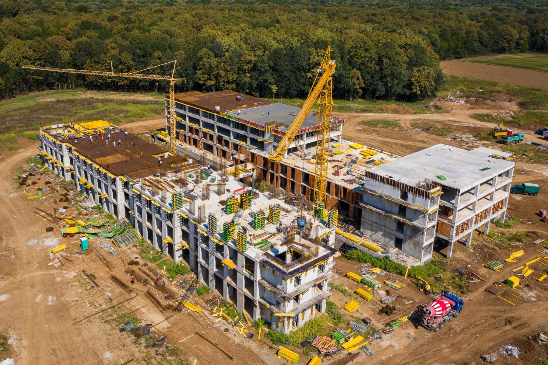 Șantier rezidențial fotografiat din dronă — fază de structură, macara activă, complex situat la marginea pădurii.