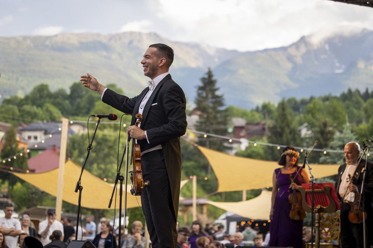 Dirijorul-violonist Bogdan Costache, în frac, cu vioara în mână, pe scena în aer liber din Borșa, cu Munții Rodnei în fundal.
