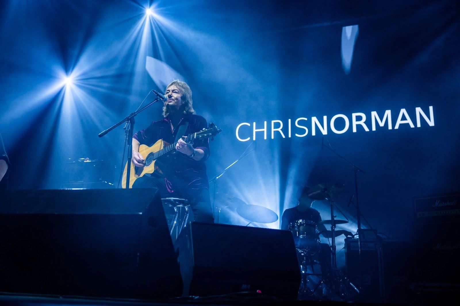 Chris Norman cu chitară acustică în concert RO, sub spoturile albastre de scenă; pe ecranul de fundal, textul „CHRISNORMAN"