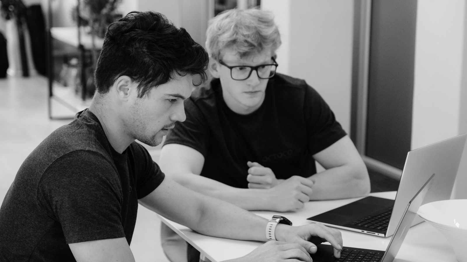 Two men sitting at a table, focused on a laptop screen, with one typing and the other watching intently. Black and white image.