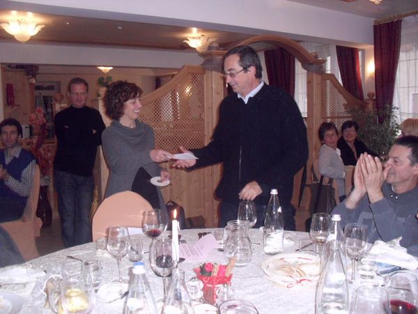 Al ristorante un uomo con gli occhiali porge una busta bianca a una donna sorridente in maglione grigio, mentre un commensale applaude