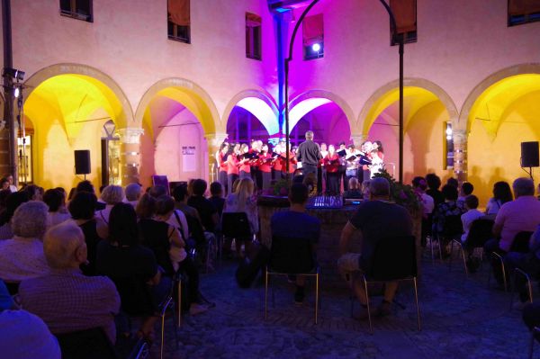 Il chiostro di sera, visto dal fondo: il pubblico seduto nel cortile e, sotto i portici illuminati di giallo e di viola, il coro che canta