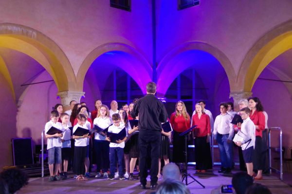 Sul palco sotto gli archi illuminati il direttore, di spalle, guida bambini in camicia bianca e adulti in camicia rossa o bianca
