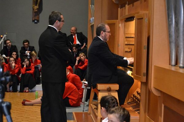 Andrea Macinanti suona alla consolle di legno dell’organo; accanto a lui, in piedi, un uomo in abito scuro, dietro i coristi seduti sui gradini