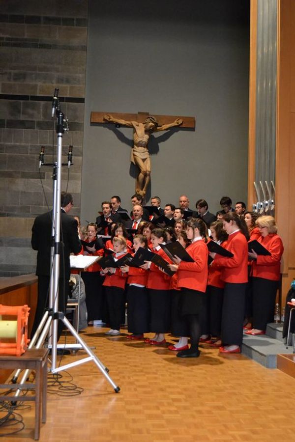 Il coro canta sotto un grande crocifisso di legno, le coriste in giacca rossa e gli uomini in abito scuro; in primo piano il direttore e i microfoni