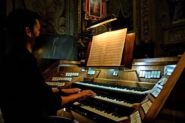 Un organista con la mascherina nera suona alla consolle a tre tastiere, con lo spartito aperto sul leggio illuminato