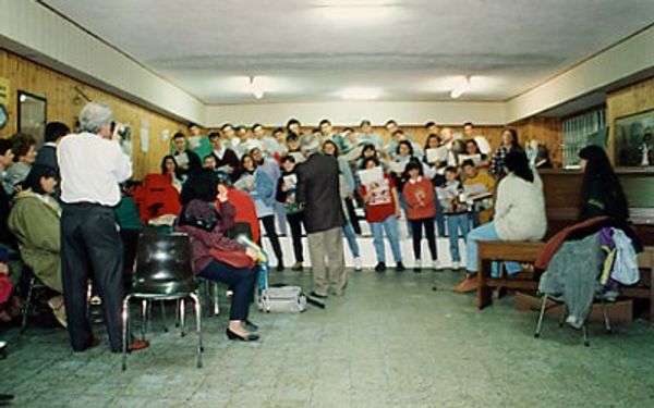 In una sala rivestita di legno il coro, in abiti di tutti i giorni, canta davanti a un piccolo pubblico; a sinistra un uomo scatta una foto
