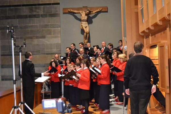 Il coro canta sotto il crocifisso di legno diretto da un uomo in abito scuro; in primo piano una videocamera, a destra la consolle dell’organo