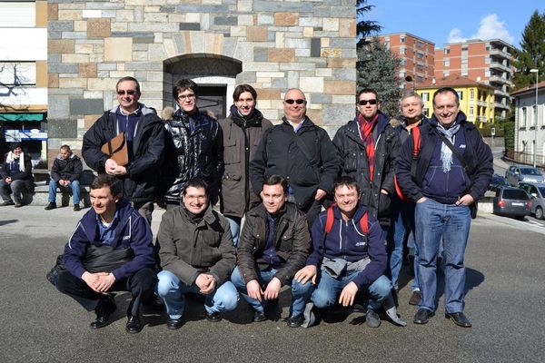 Undici uomini in giacca invernale, alcuni con gli occhiali da sole, in posa davanti a un edificio di pietra; quattro sono accovacciati in prima fila