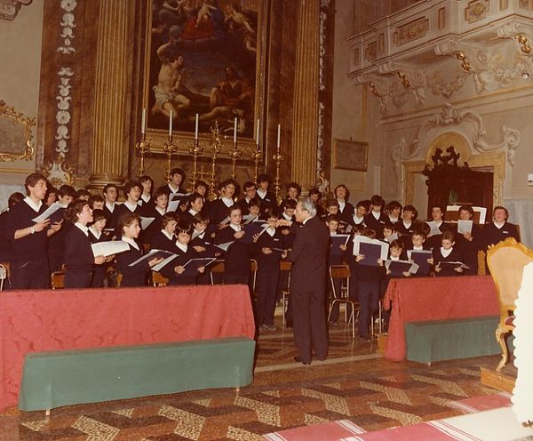 Il coro di ragazzi, in maglione blu e colletto bianco, canta con gli spartiti in mano davanti a un altare con sei candelieri, di fronte al direttore
