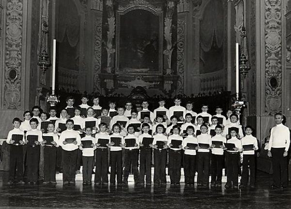 Foto in bianco e nero: una quarantina di ragazzi in dolcevita bianco, con le cartelline in mano, davanti all’altare; a destra un uomo vestito come loro