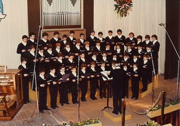 Il coro di ragazzi, in divisa scura con il colletto bianco, canta su un palco davanti alle canne dell’organo, guidato dal direttore di spalle
