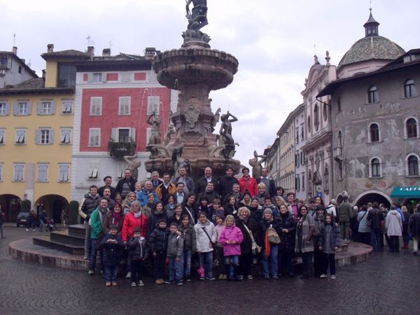Foto di gruppo della comitiva, adulti e bambini, davanti a una grande fontana con statue in una piazza tra case colorate e affrescate