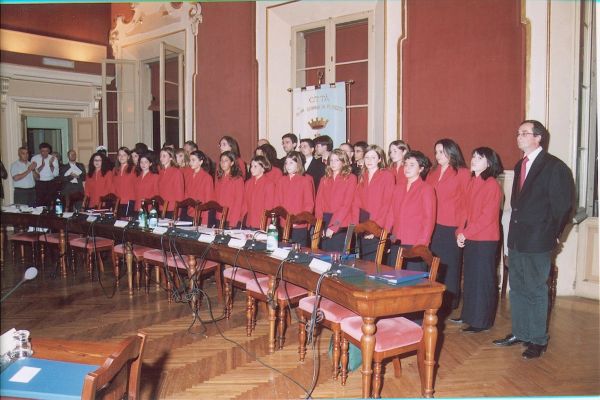 Il coro in piedi dietro il lungo banco con i microfoni, le donne in giacca rossa e gli uomini in abito scuro, sotto il gonfalone della città