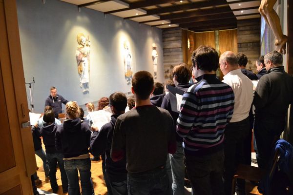 Prova in una cappella con statue di santi sulle pareti azzurre: i coristi, visti di spalle, leggono gli spartiti diretti da un uomo in fondo