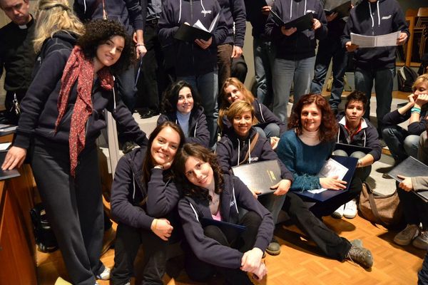 Coriste in felpa blu sedute sul parquet e sui gradini, sorridenti, con le cartelline in mano; dietro di loro, in piedi, altri coristi