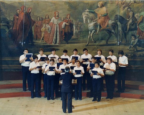 Una ventina di ragazzi in camicia bianca cantano davanti a un grande dipinto con dignitari e cavalieri; in primo piano il direttore di spalle