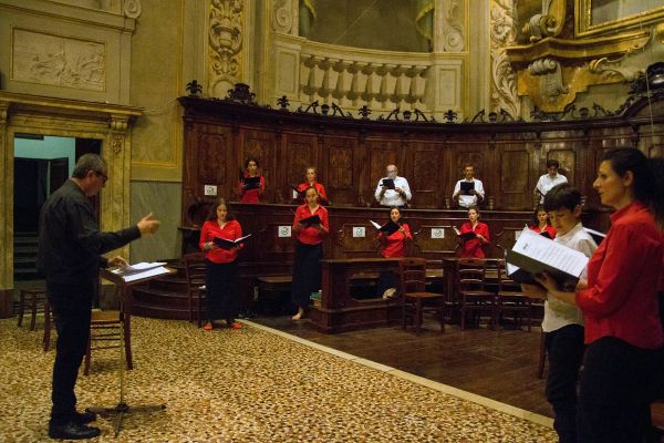 Il coro canta distanziato tra gli stalli di legno del presbiterio, le donne in camicia rossa e gli uomini in bianco; a sinistra il direttore al leggio