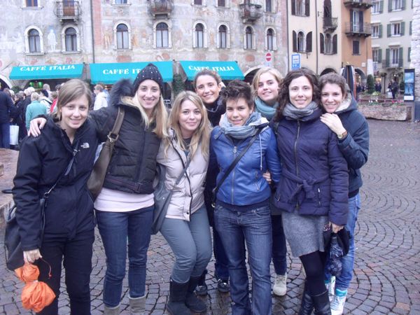Otto ragazze abbracciate in posa in una piazza lastricata, davanti alle facciate affrescate e alle tende verdi del Caffè Italia