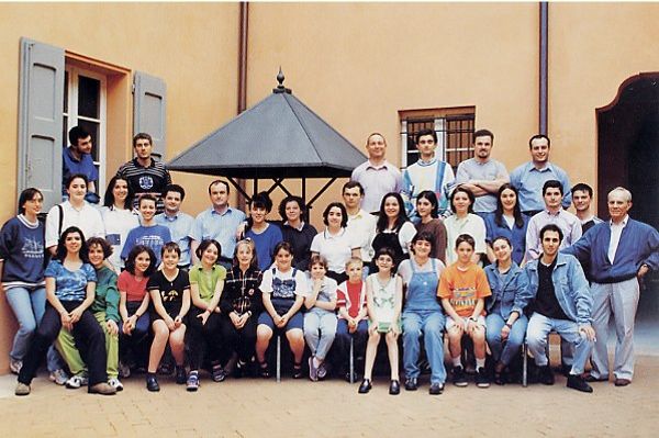 Foto di gruppo in un cortile color ocra con una tettoia a piramide: una quarantina tra ragazze, ragazzi e adulti in abiti estivi