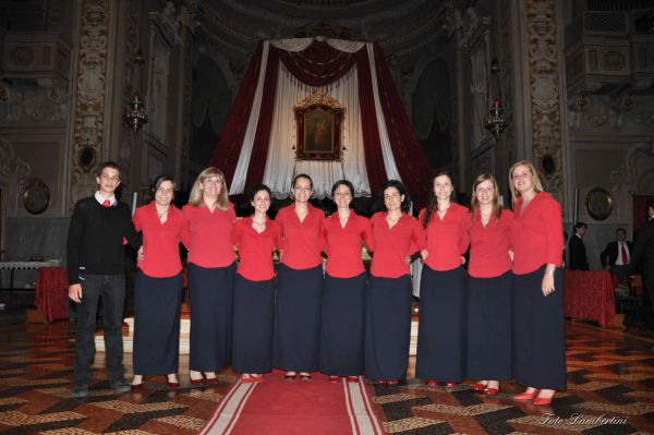 Nove coriste in camicia rossa e gonna lunga, con un ragazzo in maglione nero, in fila davanti all’altare