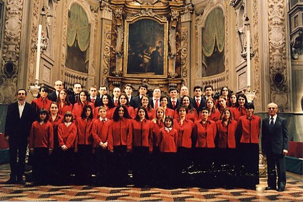 Foto di gruppo davanti all’altare: ragazze e donne in giacca rossa, dietro gli uomini in abito scuro e cravatta rossa; ai lati due uomini in completo