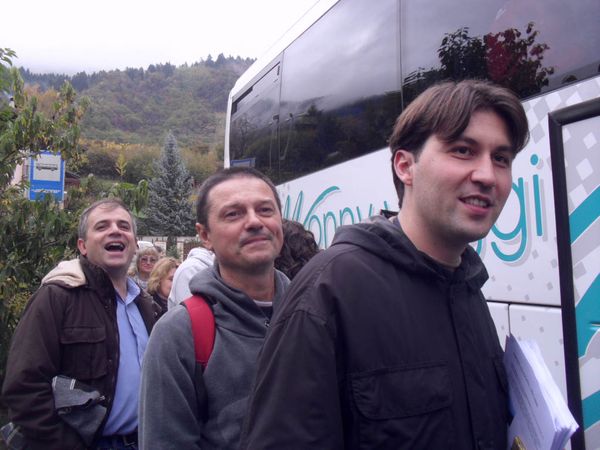 Tre uomini in fila accanto a un pullman bianco, con i monti boscosi sullo sfondo: uno ride, il più giovane tiene in mano dei fogli