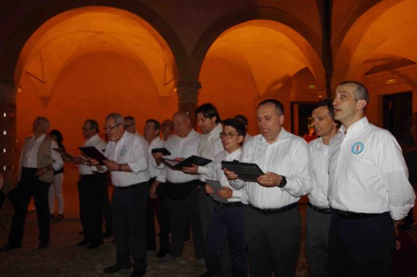 Uomini e un ragazzo in camicia bianca con uno stemma sul petto cantano leggendo le cartelline, sotto le volte del portico illuminate di arancione