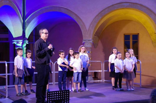 Sul palco, sotto i portici illuminati di blu e di giallo, un uomo in camicia nera parla al microfono; dietro, una decina di bambini in polo bianca