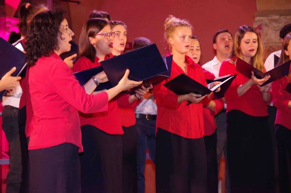 Primo piano delle coriste in camicia rossa e gonna nera che cantano leggendo le cartelline; dietro di loro alcuni uomini in camicia bianca