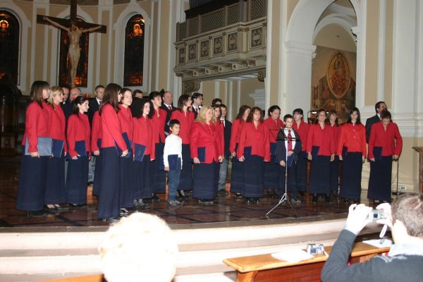 Il coro in piedi sui gradini del presbiterio sotto un grande crocifisso, le donne in giacca rossa e gonna blu; in primo piano uno spettatore fotografa