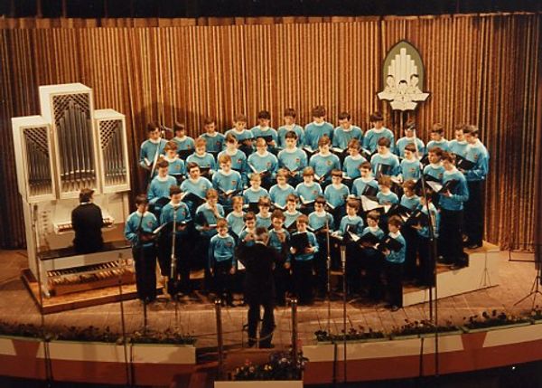 Una cinquantina di ragazzi in maglia azzurra cantano su un palco dal sipario dorato accanto a un piccolo organo bianco, diretti da un uomo di spalle