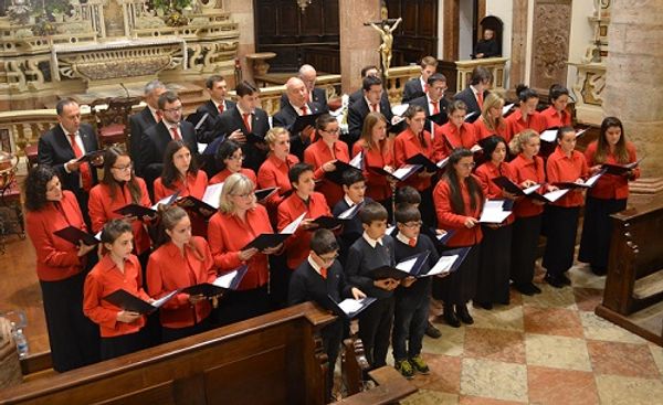 Il coro canta in una chiesa, visto dall’alto: le donne in rosso, gli uomini in abito scuro, i bambini in maglione blu e cravatta rossa