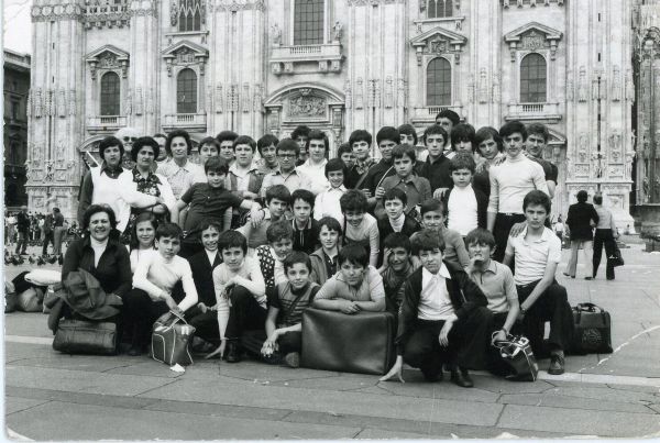 Foto in bianco e nero: una quarantina di ragazzi e alcune donne in posa sulla piazza, con valigie e borse ai piedi, davanti alla facciata gotica