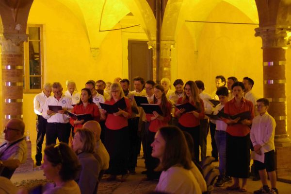 Sotto il portico illuminato di giallo, donne in camicia rossa e uomini in camicia bianca cantano insieme; in primo piano il pubblico seduto