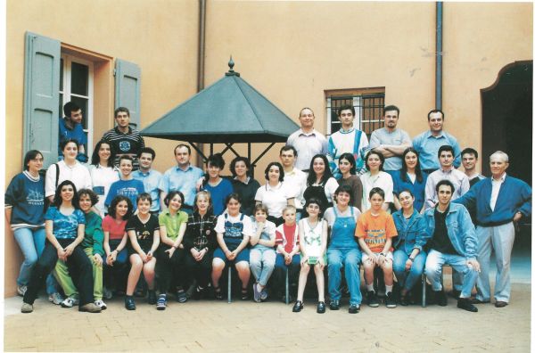 Una quarantina tra ragazze, ragazzi e adulti in abiti estivi in un cortile color ocra con una tettoia a piramide; a destra un uomo dai capelli bianchi