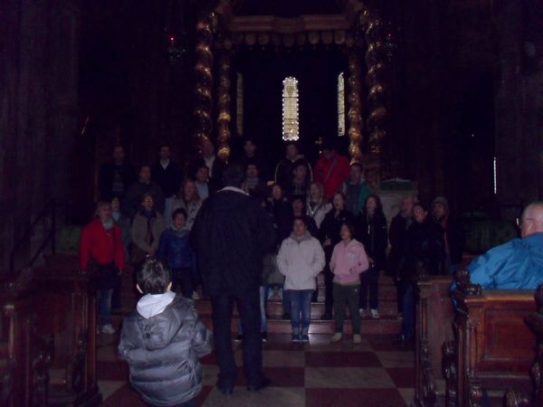 In una chiesa in penombra il coro, in abiti invernali, canta sui gradini sotto un baldacchino a colonne tortili dorate, diretto da un uomo di spalle