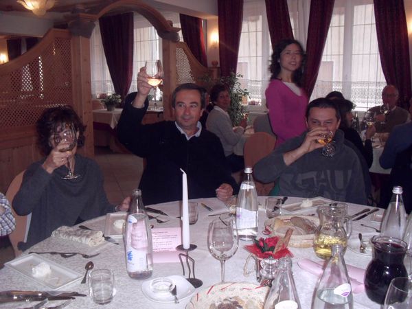 Al ristorante un uomo alza il bicchiere di vino bianco per un brindisi; accanto a lui due commensali bevono, dietro una ragazza in maglia rosa