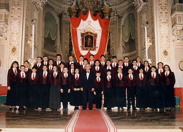 Foto di gruppo davanti all’altare con un drappo rosso e bianco: ragazze e ragazzi in giacca bordeaux e cravatta rossa, al centro un uomo in smoking