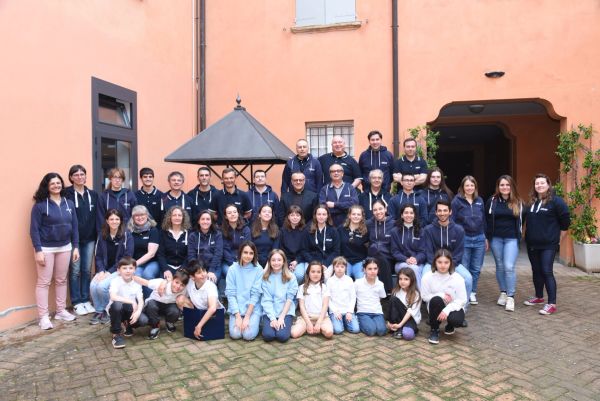 Foto di gruppo in un cortile color salmone con una tettoia a piramide: adulti e ragazzi in felpa blu, davanti i bambini seduti a terra