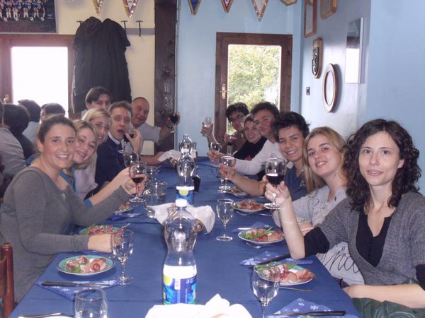 Una lunga tavolata con la tovaglia blu: i commensali, quasi tutti giovani, alzano i calici di vino verso l’obiettivo davanti a piatti di affettati