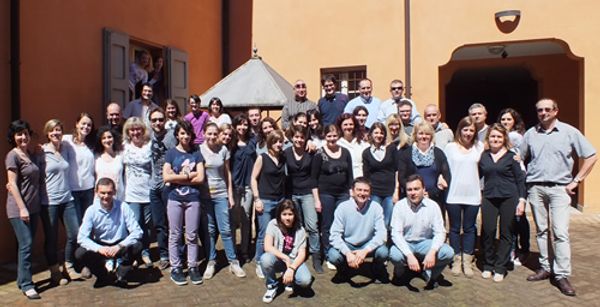 Una quarantina di coristi in abiti primaverili in posa in un cortile color ocra con una tettoia a piramide; alcuni si affacciano da una finestra